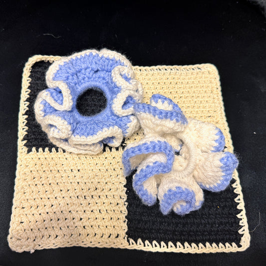 Two handmade crochet hair scrunchies in a blue and cream colorway, displayed on top of a matching cream and black crochet square on a black background.