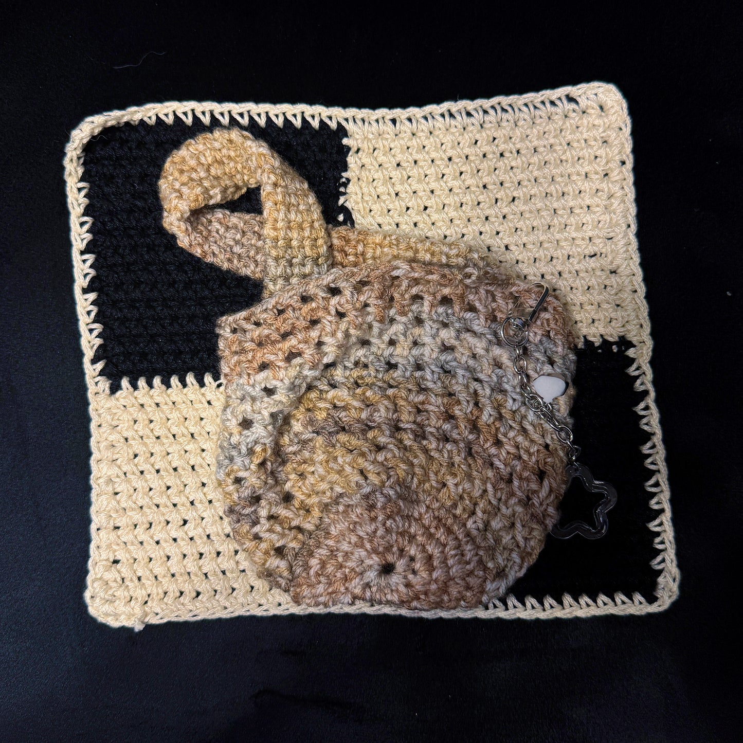 A handmade crocheted pouch with a loop handle, resting on a matching square crochet mat. The pouch features a variegated pattern of tan, brown, and beige yarn, and is photographed against a solid black background.