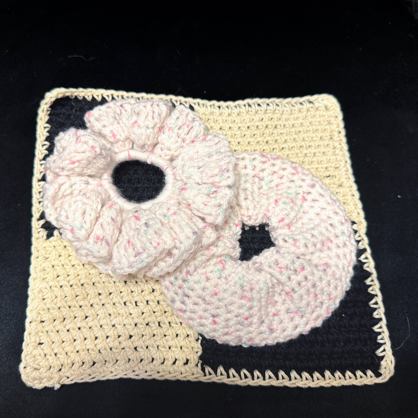 A handmade crocheted set featuring a cream-colored square dishcloth with a black printed design and two matching crocheted donut-shaped washcloths in cream with pink and green flecks, displayed on a black background.