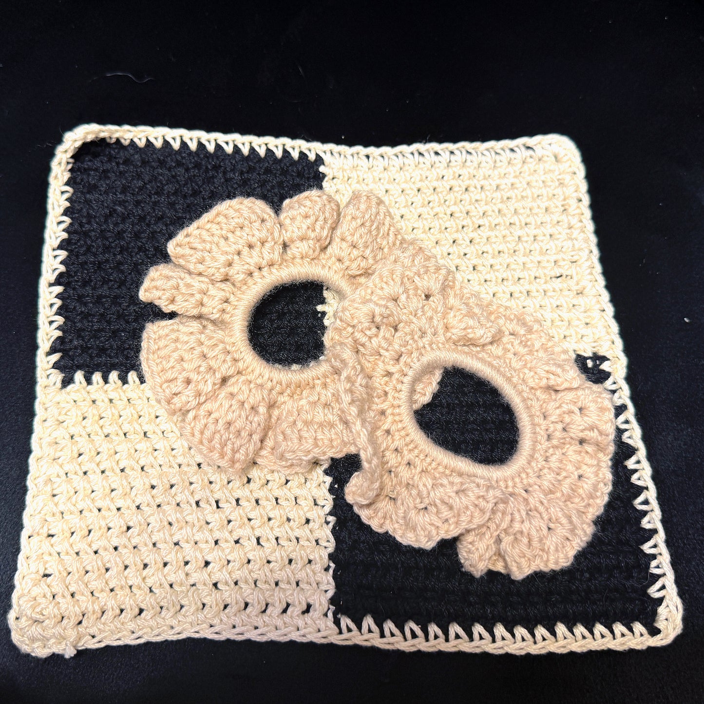 Two beige crochet hair scrunchies displayed on a black and beige checkered crochet mat against a black background.