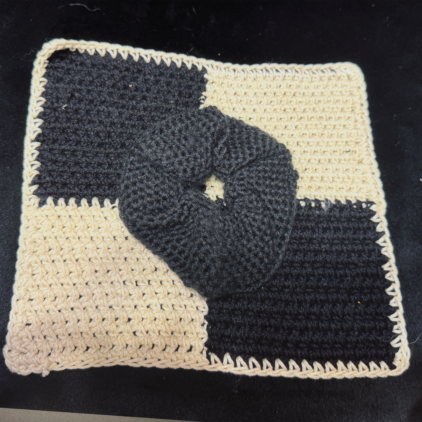 A black crocheted hair scrunchie placed on top of a black and cream checkered crochet mat, set against a dark black background.