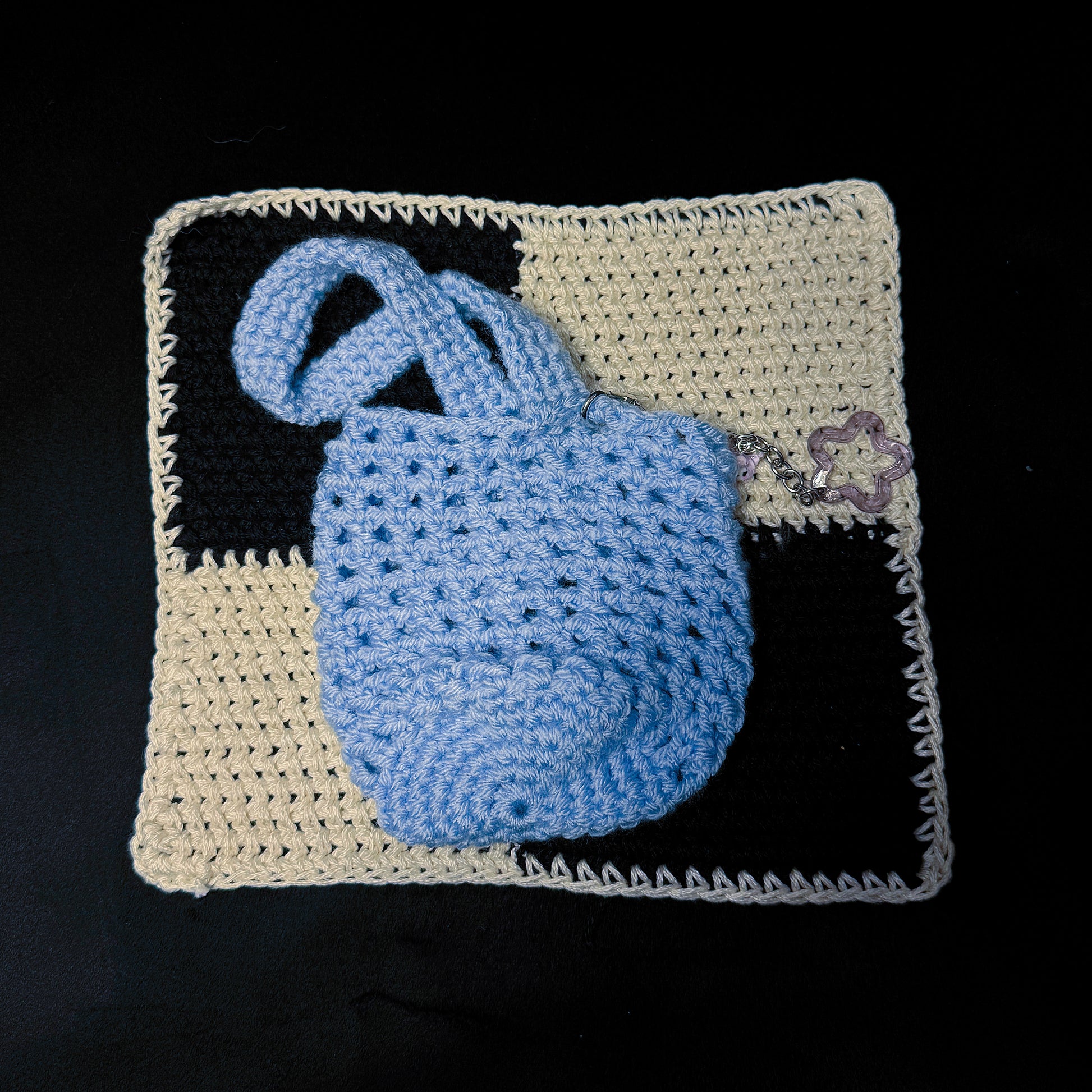 A light blue crocheted handbag with a matching top handle, displayed on a cream and black checkered crochet mat against a solid black background.