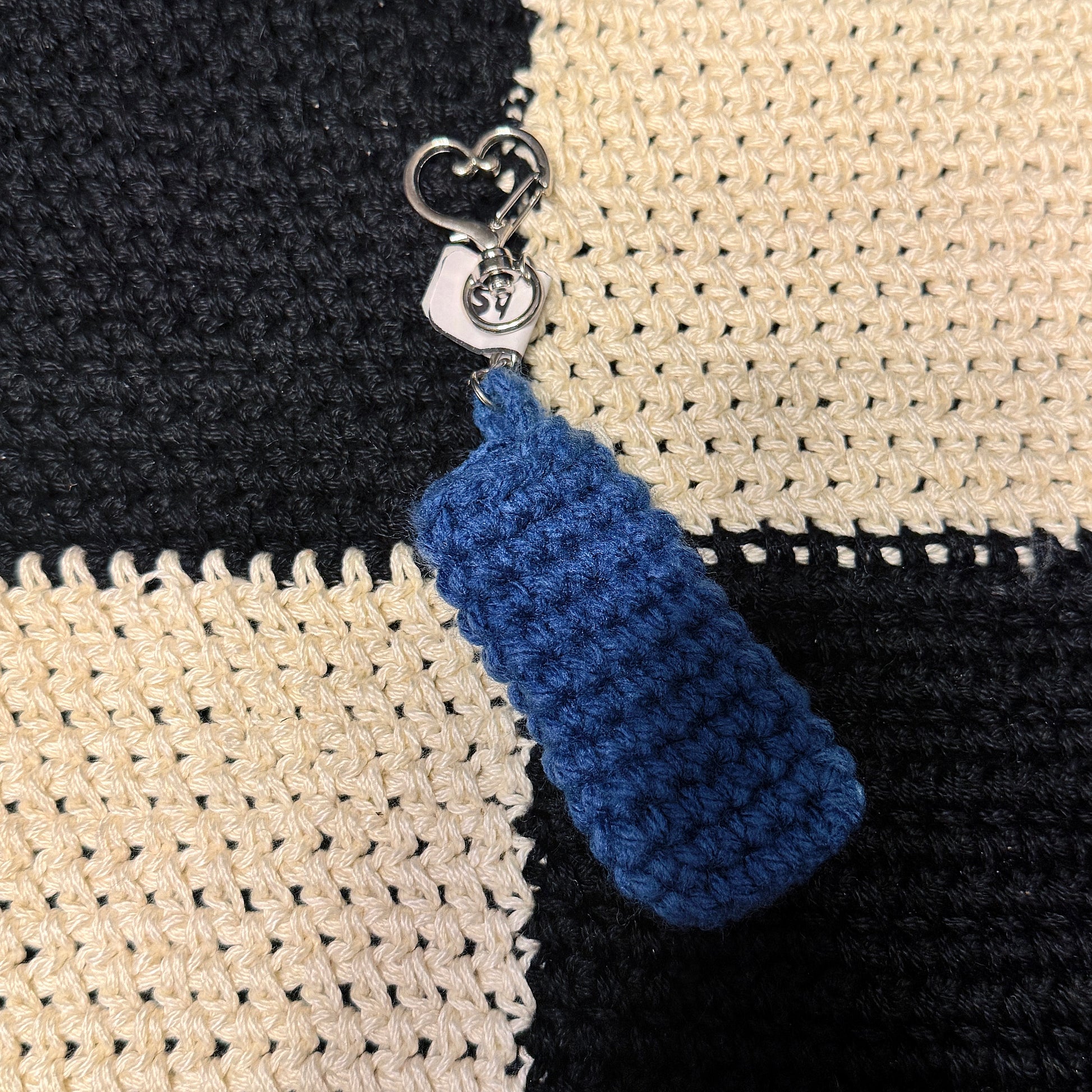 A close-up of a crocheted blue keychain or zipper pull with a silver metal clasp, shown resting on a black and cream checkered knitted fabric background.