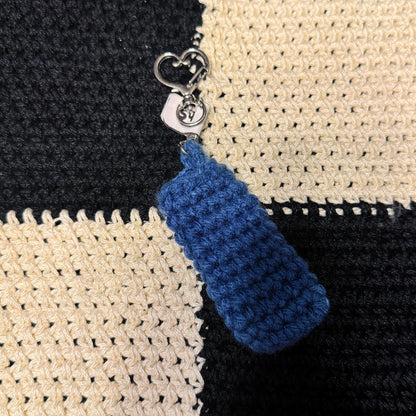 A close-up of a crocheted blue keychain or zipper pull with a silver metal clasp, shown resting on a black and cream checkered knitted fabric background.