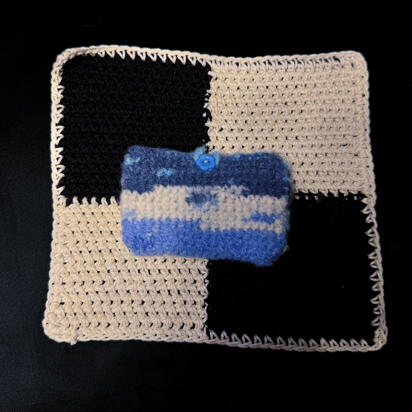 A handmade crocheted pouch with a blue and cream checkered pattern, featuring a small blue button closure, displayed on top of a larger matching crocheted mat against a black background.