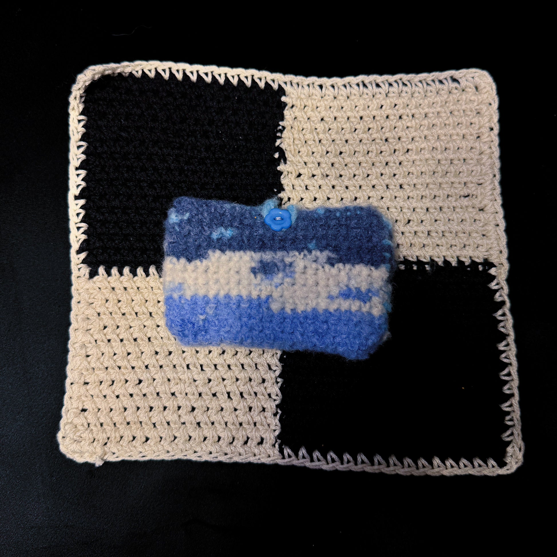 A handmade crocheted pouch with a blue and cream checkered pattern, featuring a small blue button closure, displayed on top of a larger matching crocheted mat against a black background.