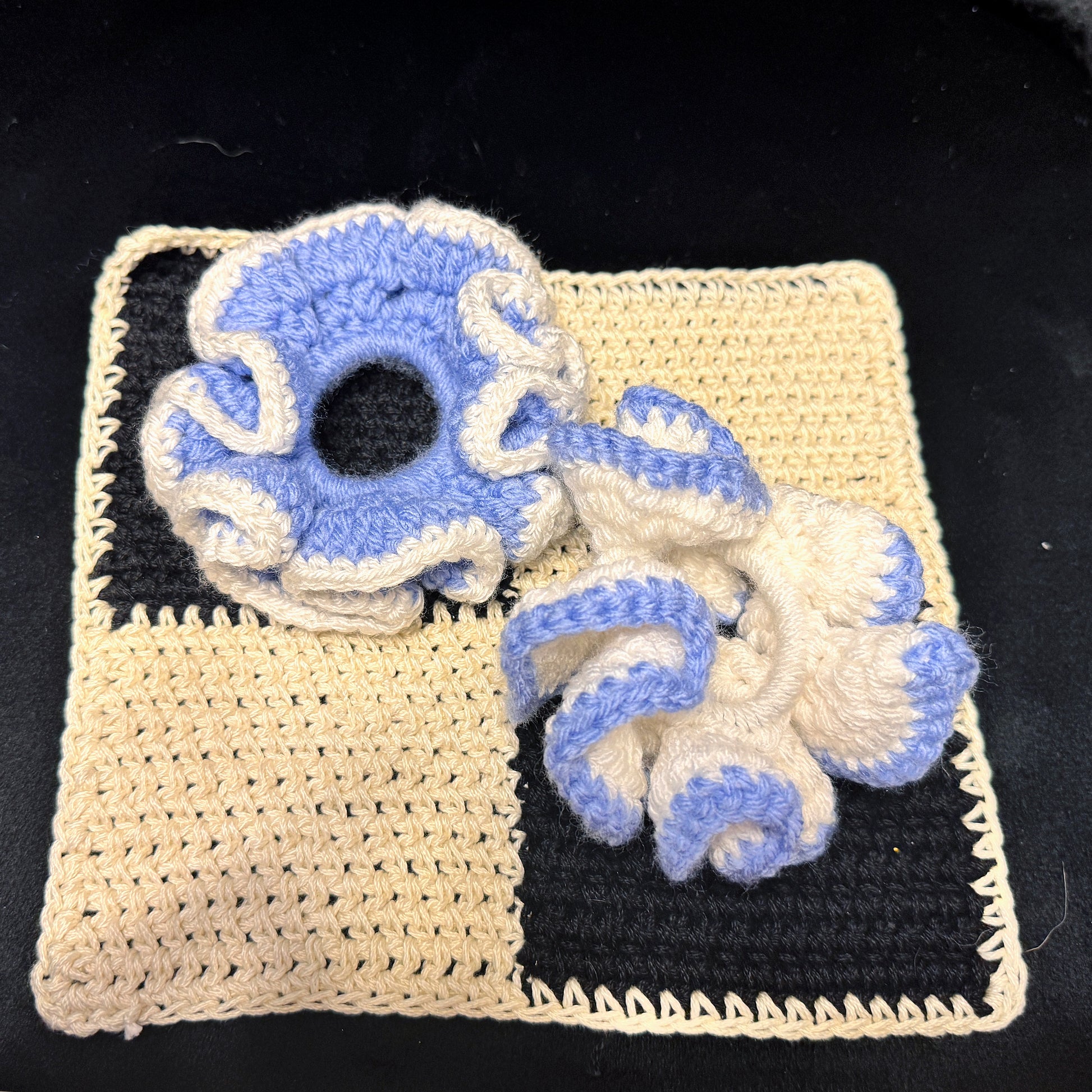 Two handmade crochet hair scrunchies in a blue and cream colorway, displayed on top of a matching cream and black crochet square on a black background.
