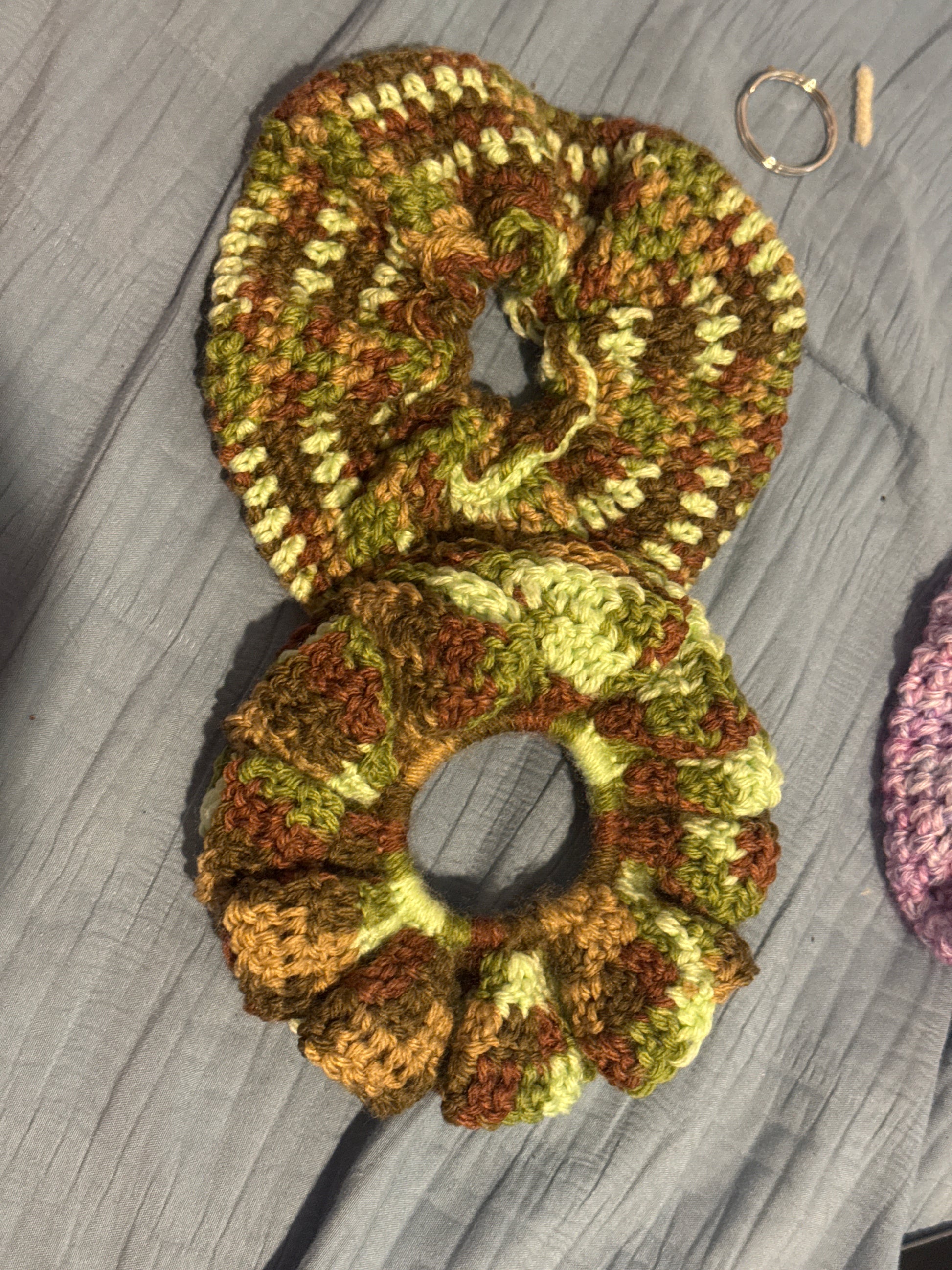 Two handmade crocheted hair scrunchies in variegated olive green, brown, and cream yarn, displayed on a gray textured fabric background.