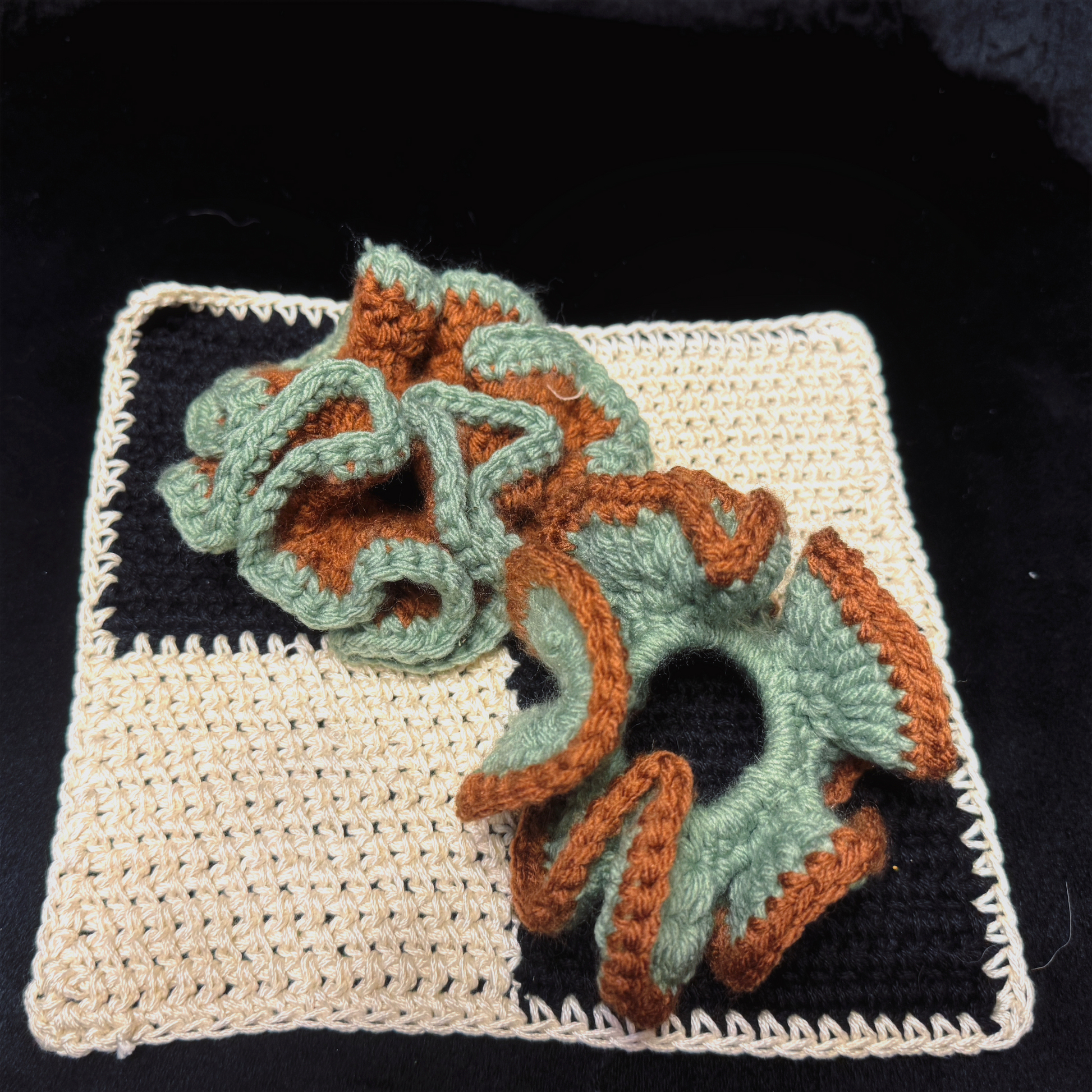 Two handmade crochet hair scrunchies in sage green and rust orange, displayed on a black and cream checkered crochet mat against a black background.