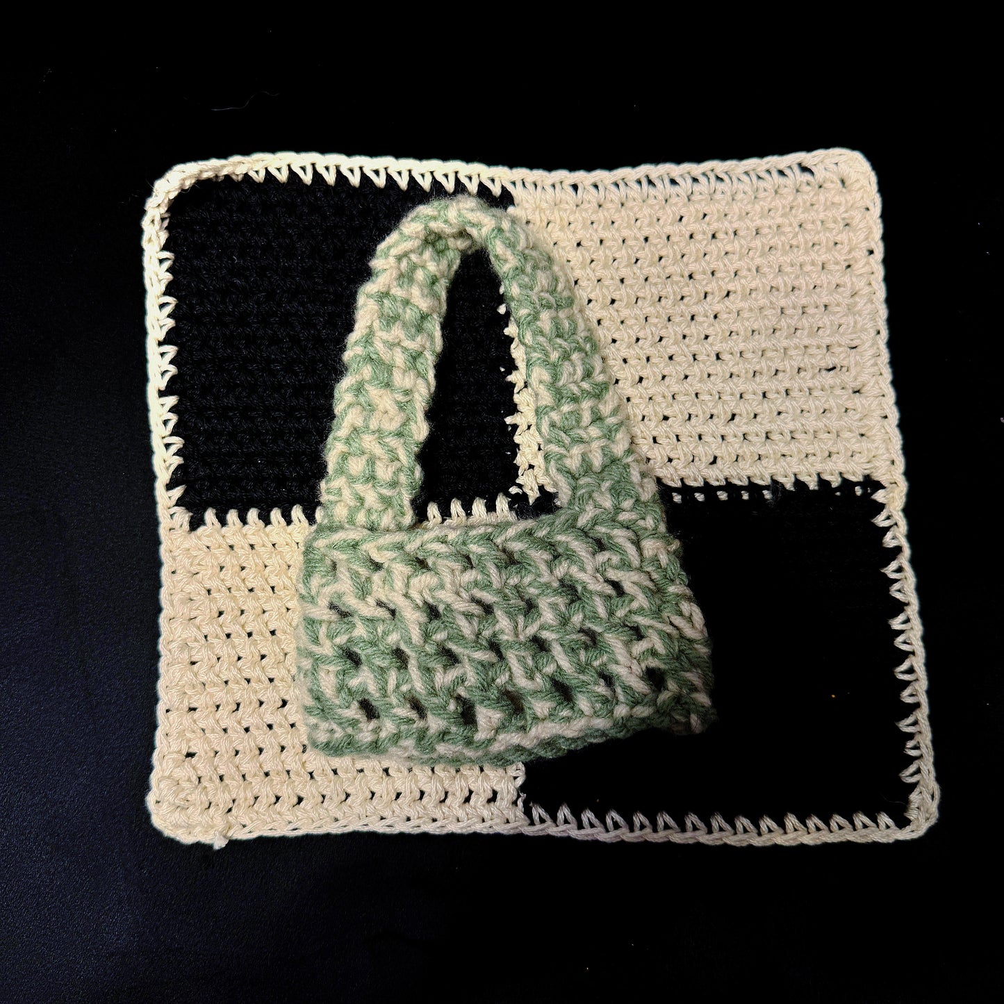 A handmade crocheted handbag in sage green and cream, displayed on a matching crocheted square mat with black and cream checkered sections against a black background.