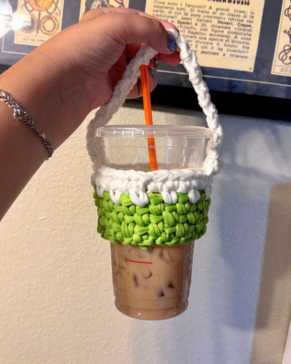 A hand holds a clear plastic cup with an iced coffee drink and an orange straw, wrapped in a handmade white and green chunky crochet sleeve with a top handle, against an indoor wall background.