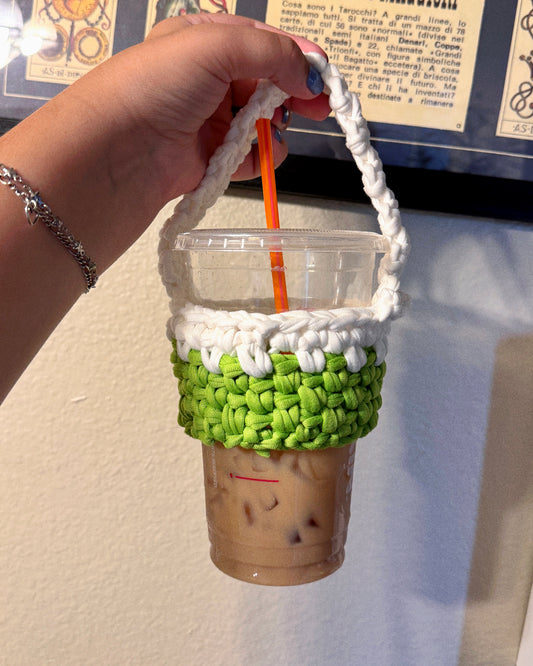 A hand holds a clear plastic cup with an iced coffee drink and an orange straw, wrapped in a handmade white and green chunky crochet sleeve with a top handle, against an indoor wall background.