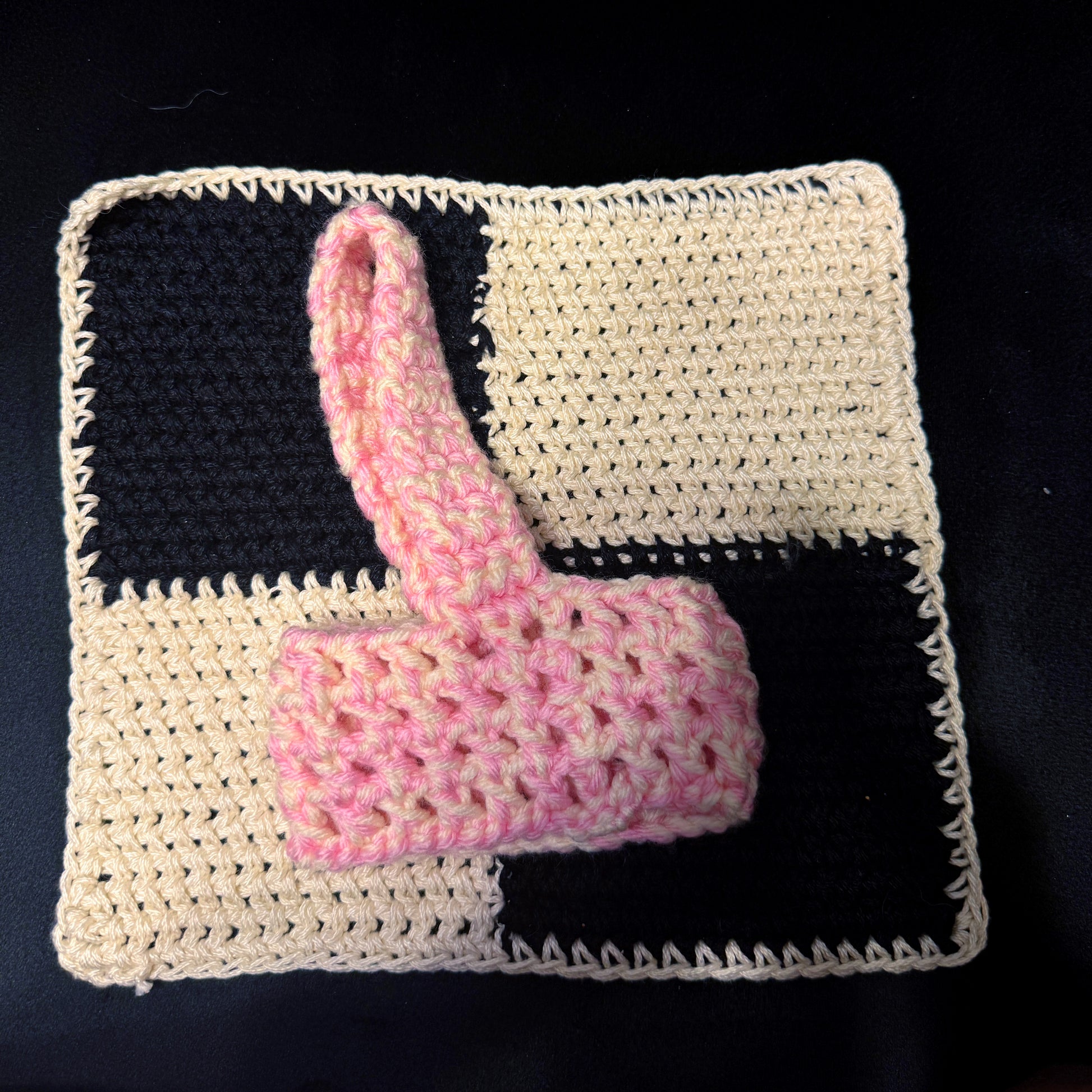 A crocheted pink and cream checkered square with a matching pink crocheted handle, displayed on a black background.