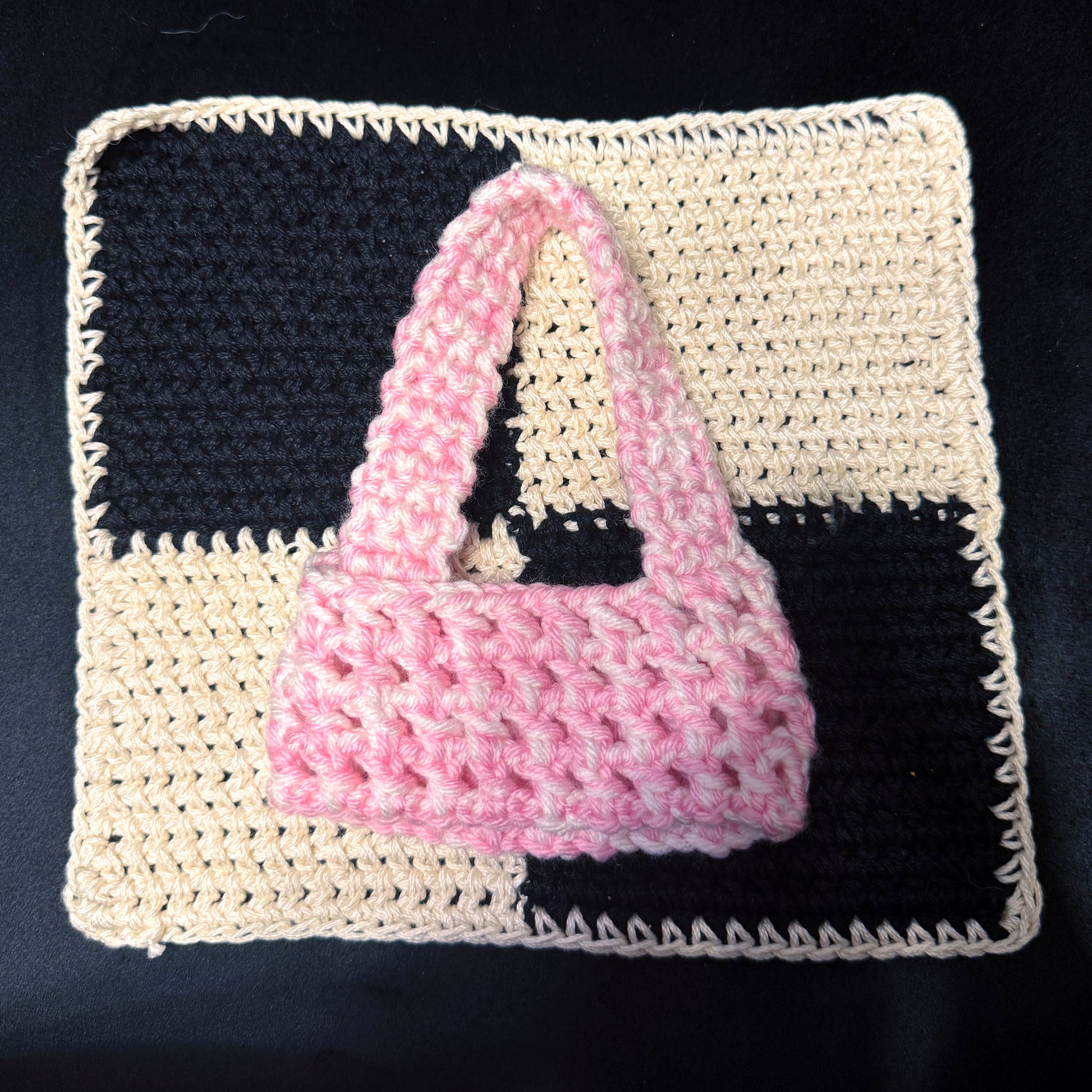 A small crocheted pink handbag with a top handle, displayed on a square cream and black checkered crochet mat against a dark background.