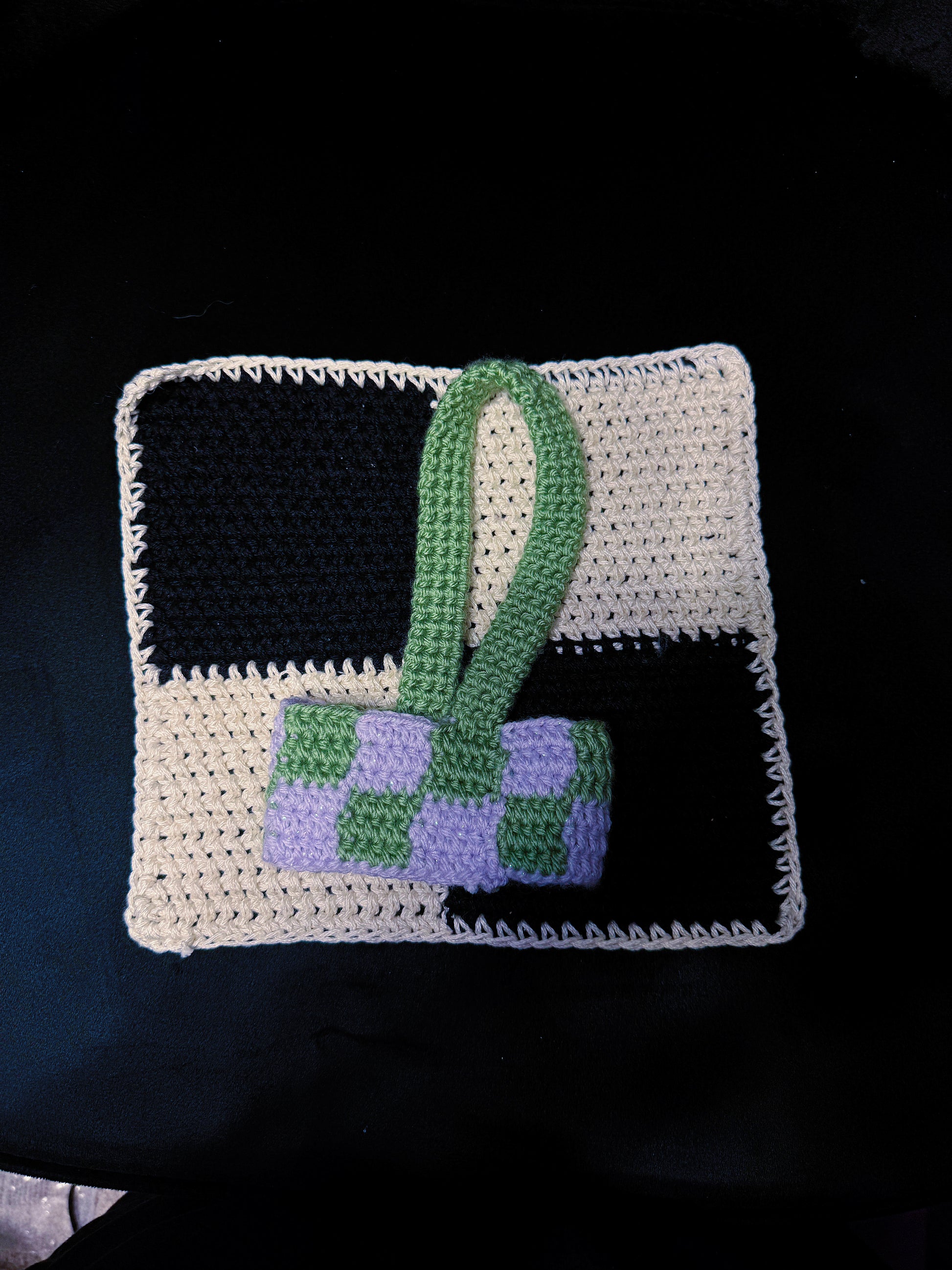 A handmade crocheted bag with a black and off-white checkered body and a green and lavender checkered front panel, featuring a green handle, displayed on a dark background.