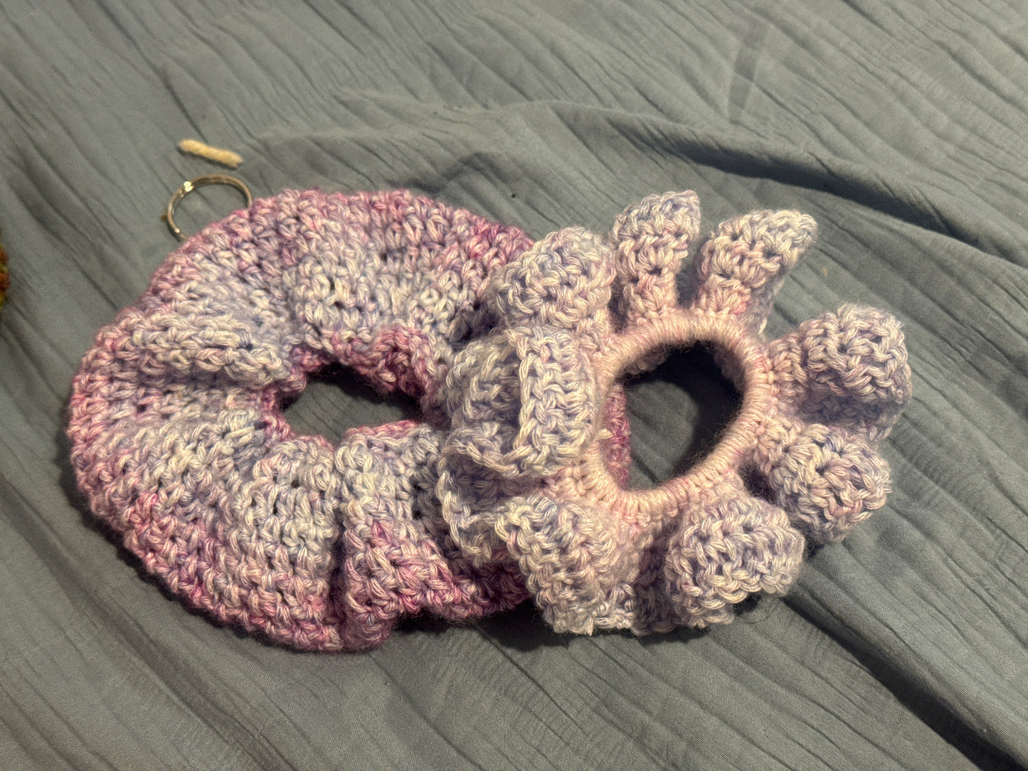 Two handmade crocheted hair scrunchies in a variegated pink, purple, and gray yarn, displayed on a textured gray fabric background.
