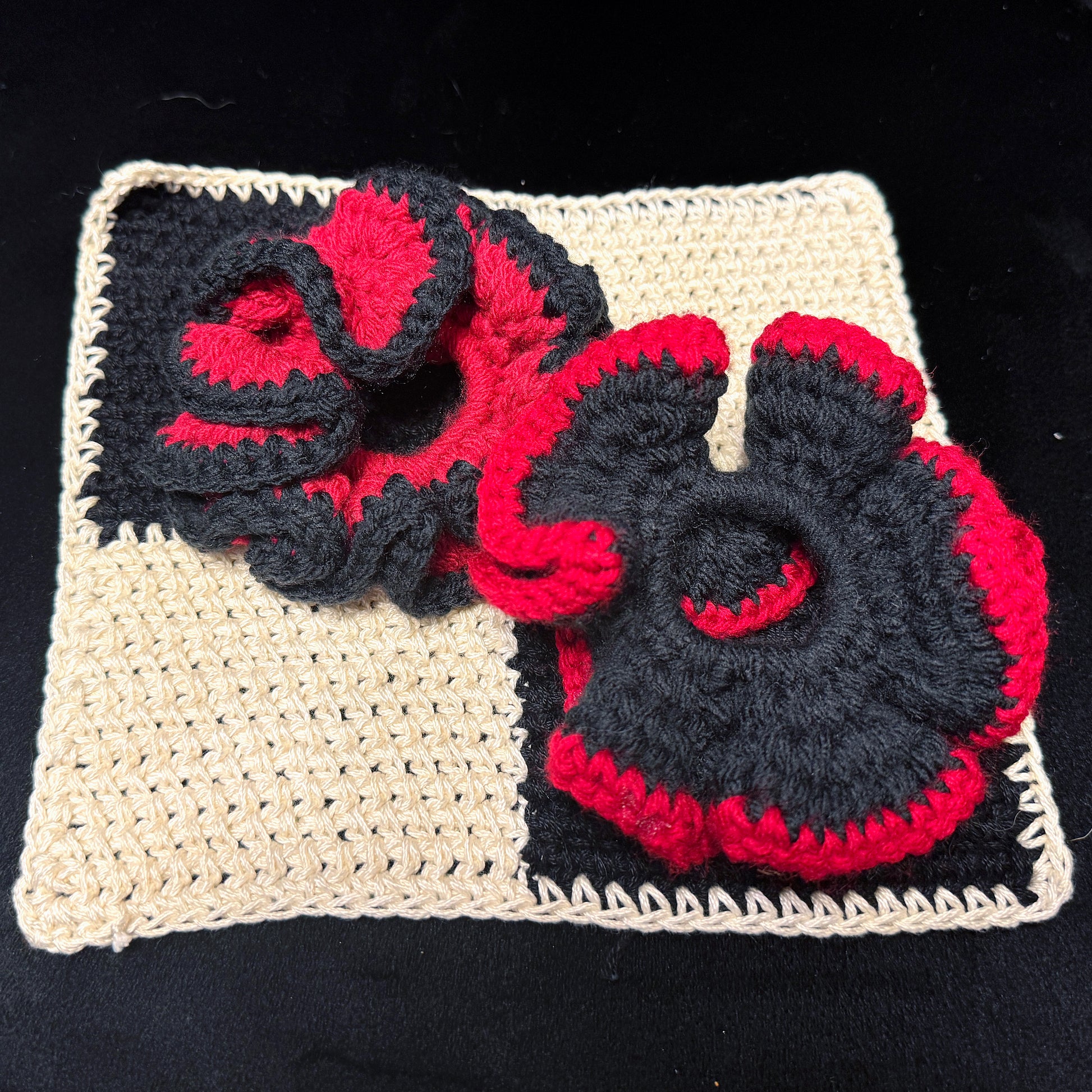 A handmade crocheted textile set featuring two large, textured poppy-like flowers in black and red yarn, arranged on top of a square cream-colored crochet mat against a black background.