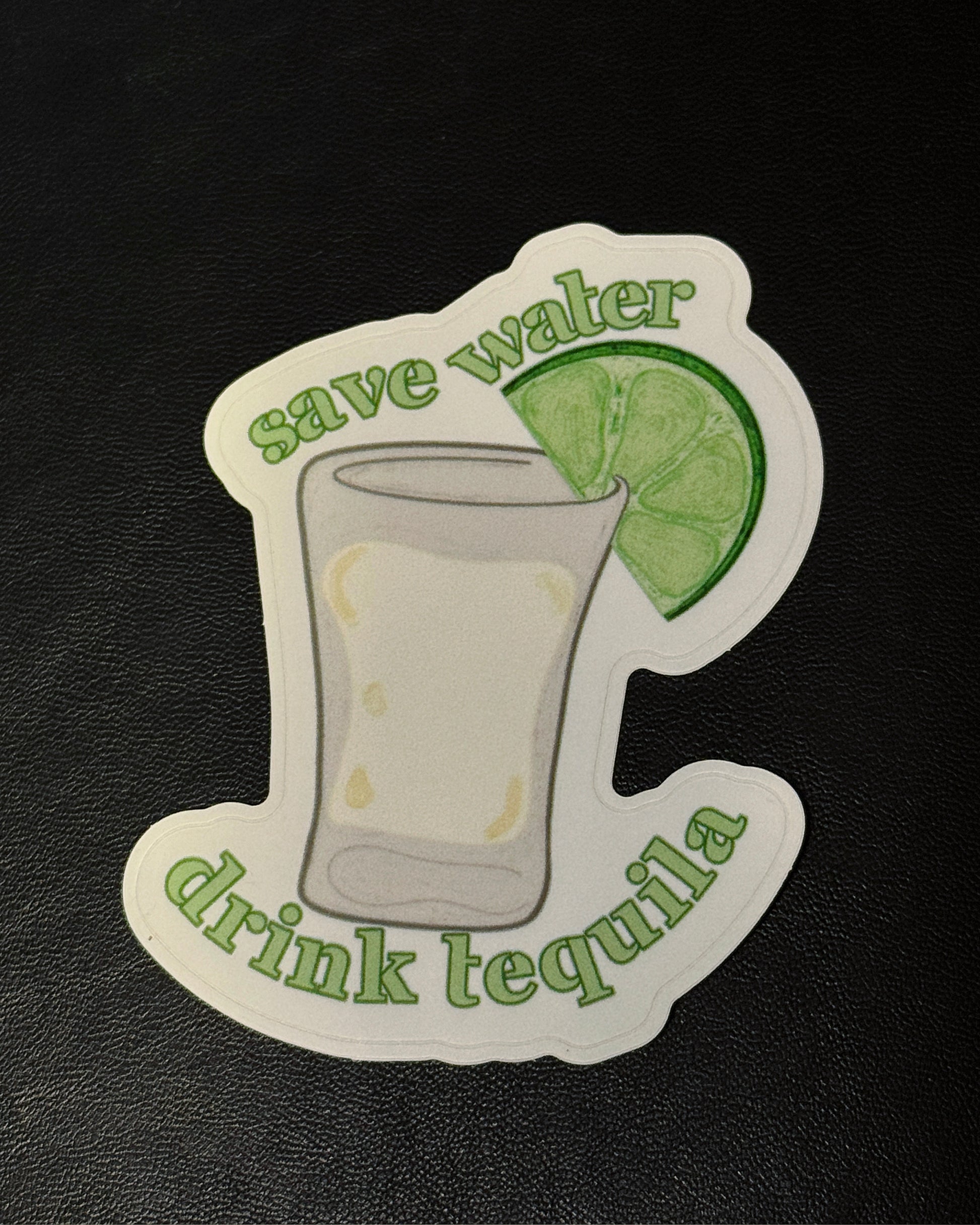 A die-cut sticker featuring an illustration of a shot glass with a lime wedge and the text 'save water drink tequila' in green, placed on a black textured surface.