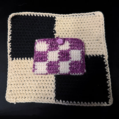 A small crocheted clutch or card holder with a purple and white checkered pattern and a purple button closure, placed on top of a larger black and cream checkered crocheted mat against a black background.